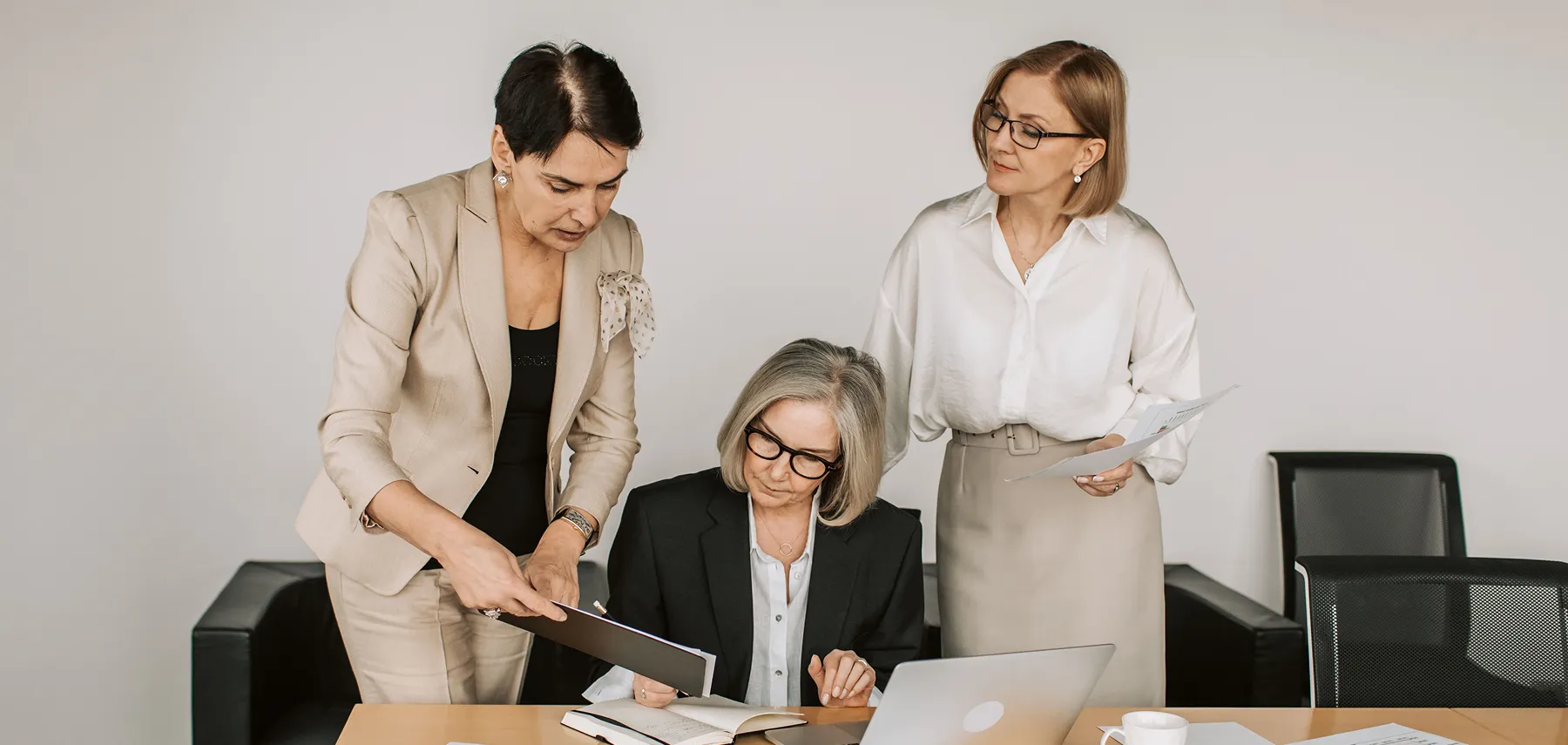 Three businesswomen reviewing documents around a laptop in an office with a beige and black decor.